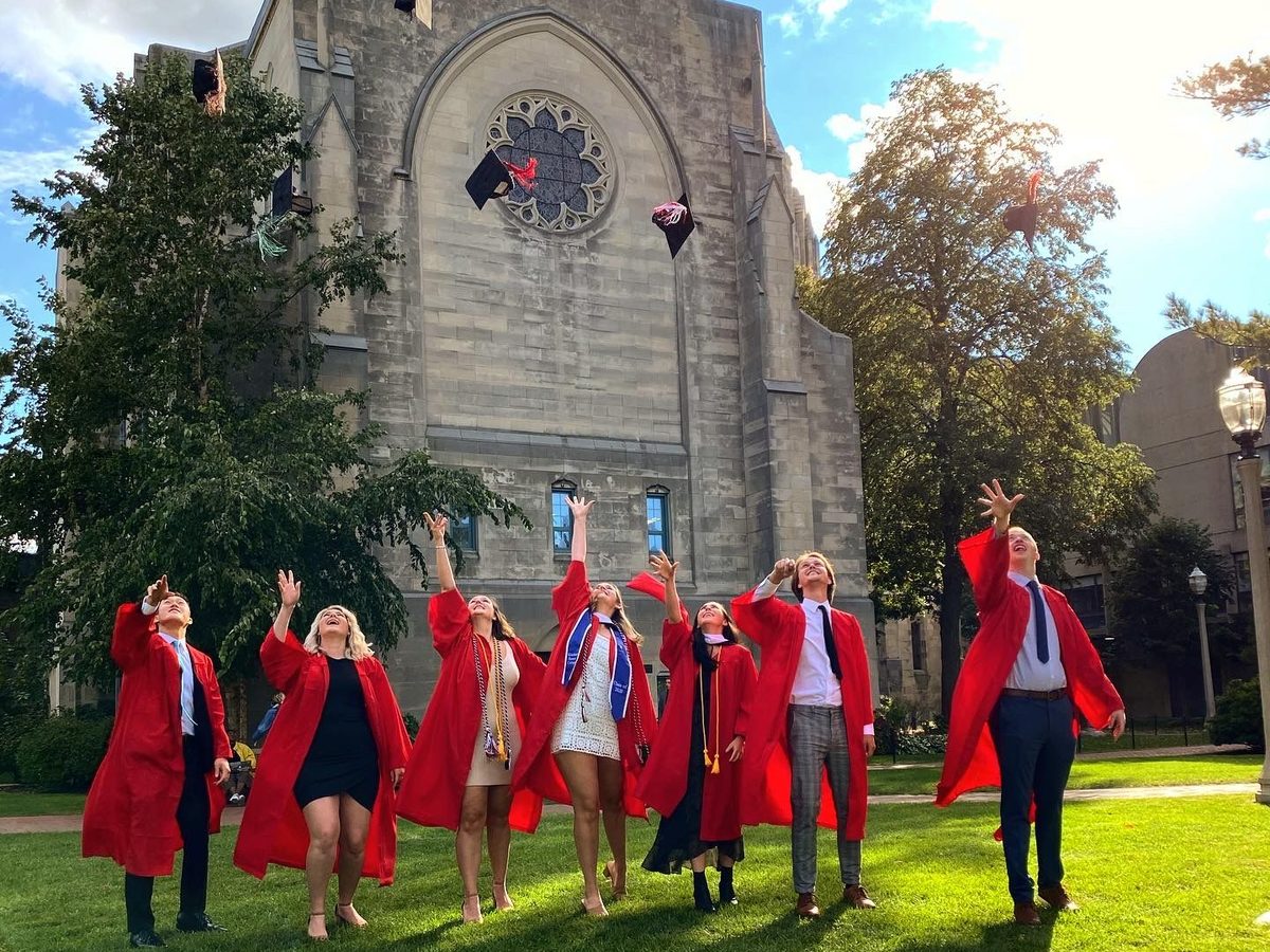Thor and six friends in red graduation gowns throwing their caps in the air in front of a stone chapel