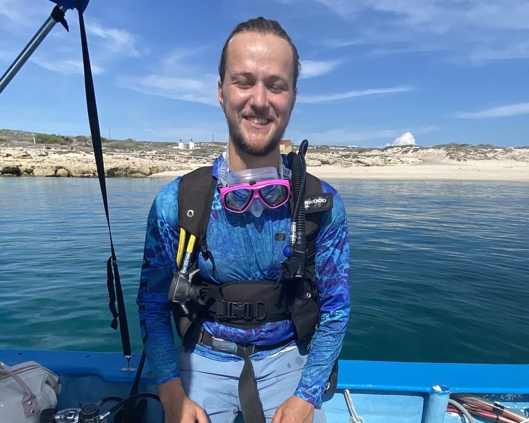 Thor on a small boat in scuba gear, smiling, blue water and a beach behind him